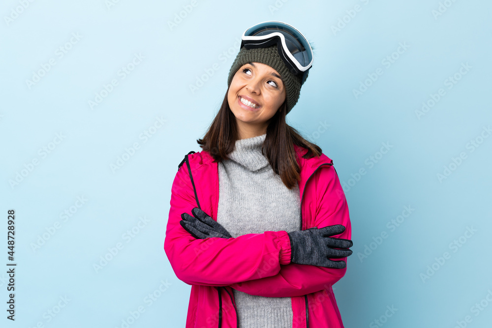 Mixed race skier girl with snowboarding glasses over isolated blue background looking up while smiling.