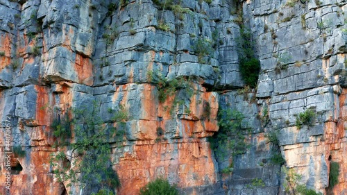 Red rugged cliff - sedimentary rock mountain formation. Vertical natural rough wall of stratum layers, colorful, sliced and cracked, brown, orange color due to ferric oxides. Geological strata canyon.