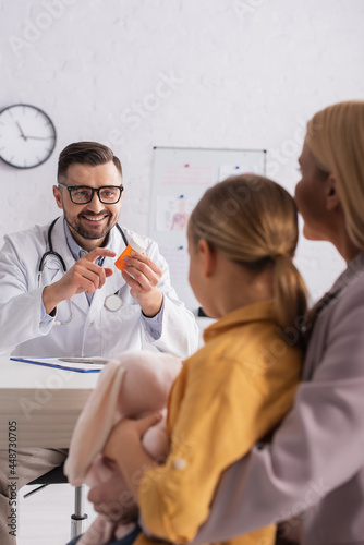 Positive pediatrician pointing at pills near blurred parent and kid