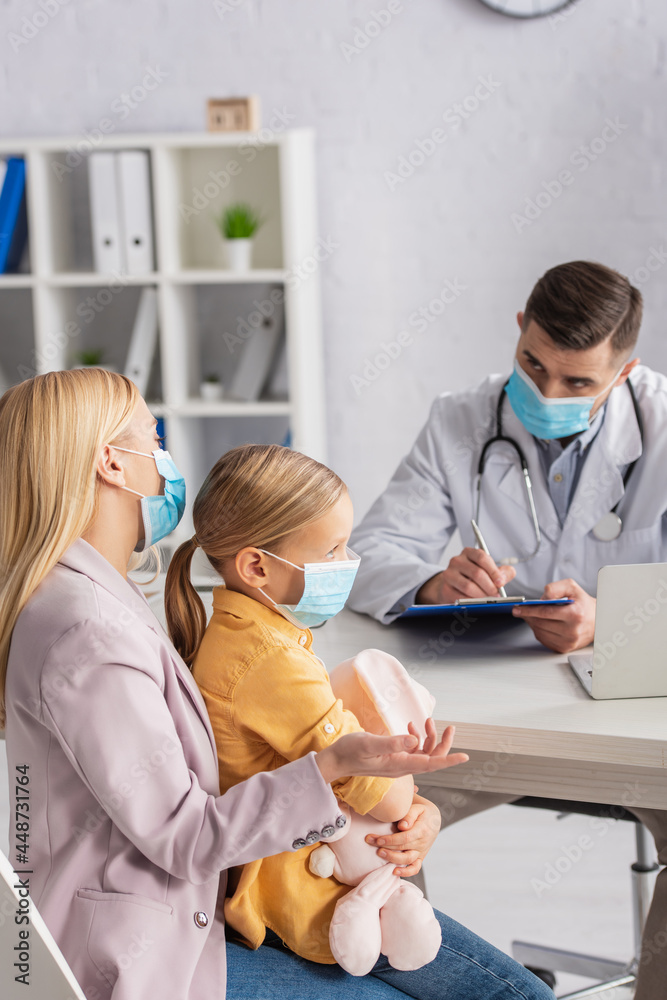 Woman hugging daughter and talking to doctor in medical mask writing on clipboard Woman hugging daughter and talking to doctor in medical mask writing on clipboard