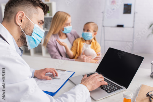 Pediatrician in medical mask using laptop near pills and family in clinic