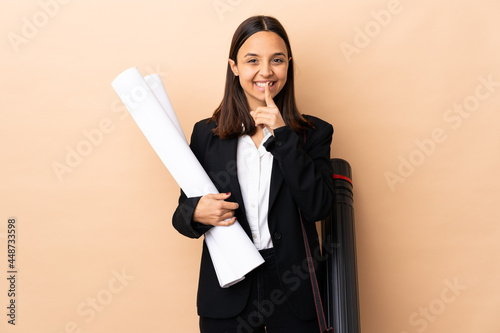 Young architect woman holding blueprints over isolated background showing a sign of silence gesture putting finger in mouth