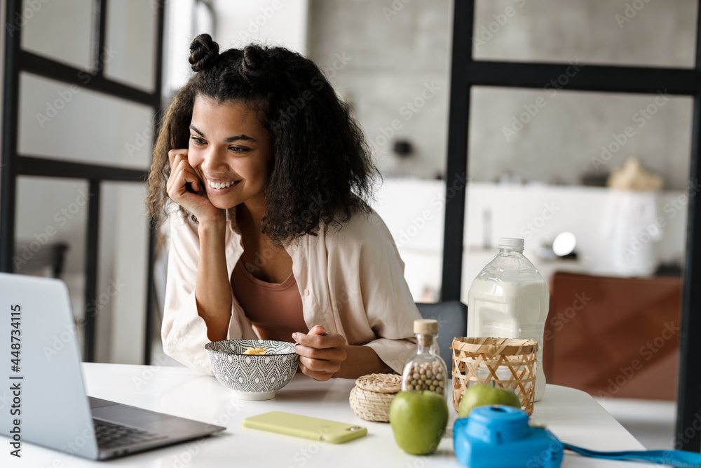 Black young woman working with laptop while having breakfast at home