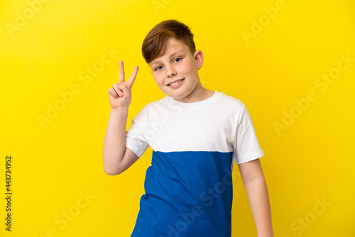 Little redhead boy isolated on yellow background smiling and showing victory sign