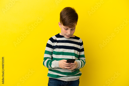 Little redhead boy isolated on yellow background sending a message with the mobile