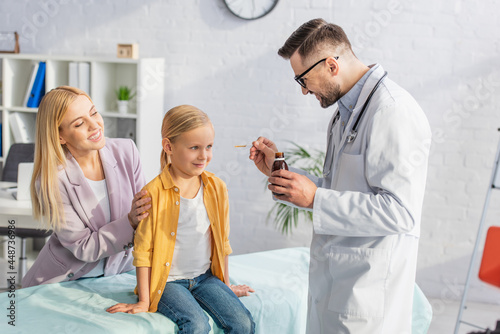 Smiling pediatrician holding syrup near smiling patient on medical couch