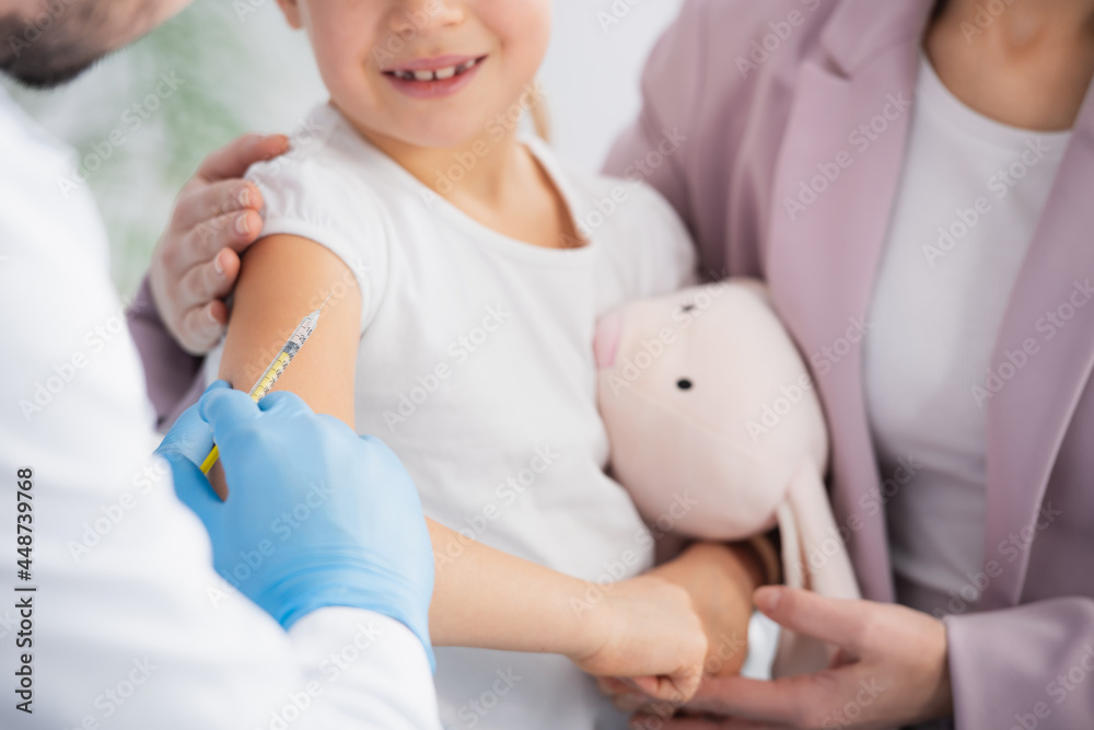 Fototapeta premium Cropped view of doctor holding syringe near smiling kid with soft toy and mother