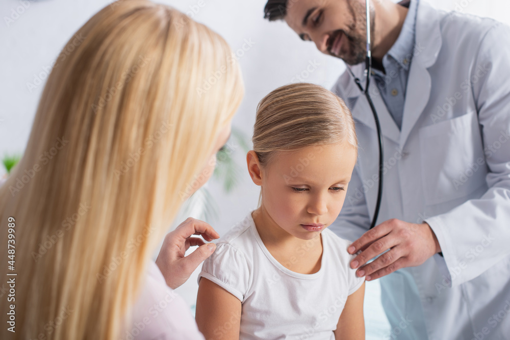 Sad kid sitting near mother and doctor with stethoscope on blurred background Sad kid sitting near mother and doctor with stethoscope on blurred background
