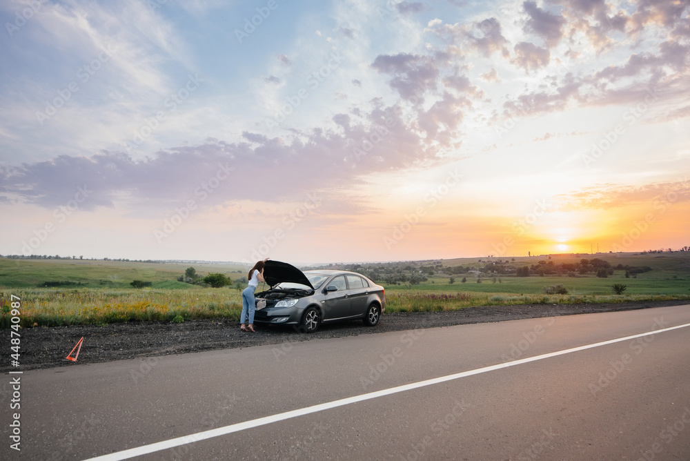 A young girl stands near a broken-down car in the middle of the highway during sunset and tries to call for help on the phone. Breakdown and repair of the car. Waiting for help. A young girl stands near a broken-down car in the middle of the highway during sunset and tries to call for help on the phone. Breakdown and repair of the car. Waiting for help.