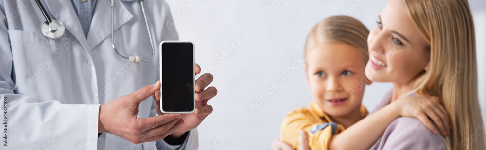 Doctor holding smartphone with blank screen near smiling mother and kid, banner