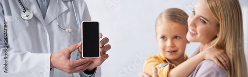 Doctor holding smartphone with blank screen near smiling mother and kid, banner