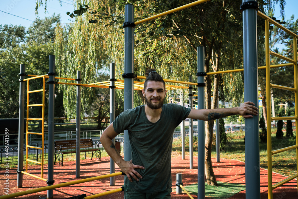 Fototapeta premium Active lifestyle concept. Young man is standing on equipped outdoor exercise area and resting. Banner with male fitness trainer. Replenishes reserve of strength and energy before strength training.