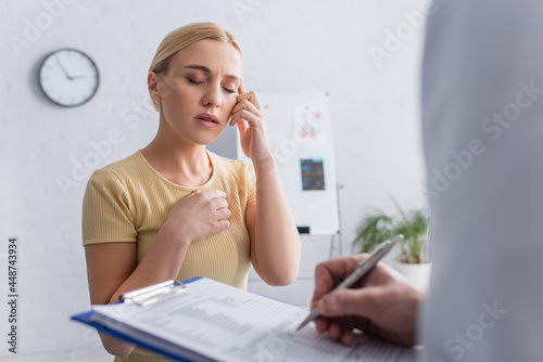 woman suffering from migraine near blurred doctor writing on clipboard