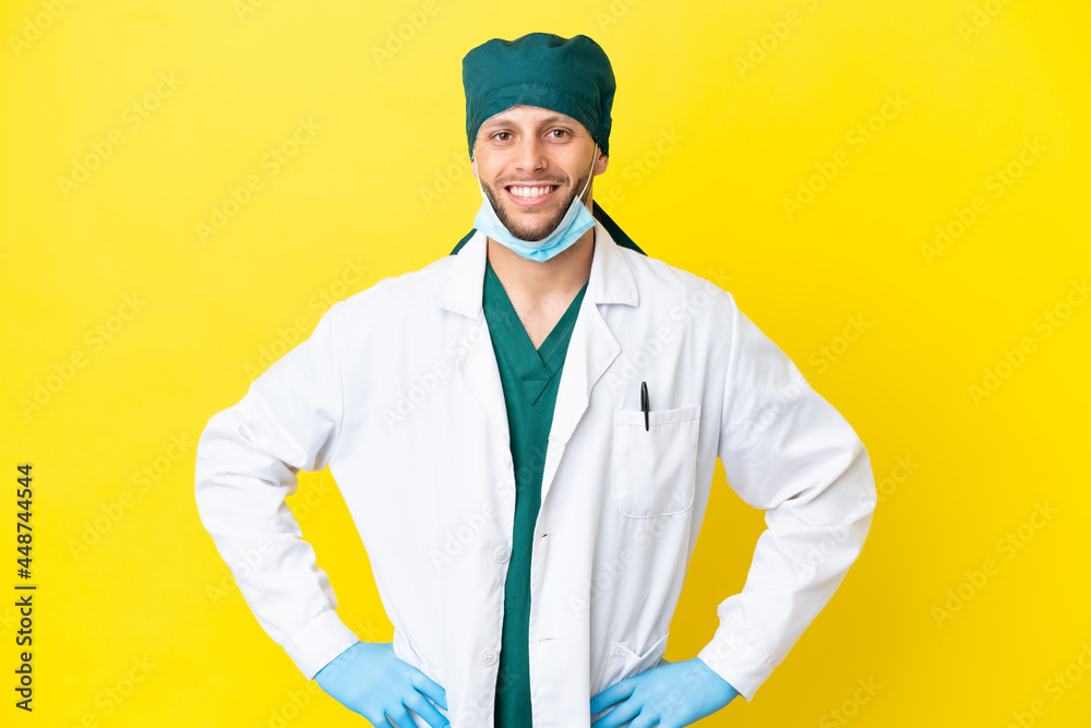 Surgeon blonde man in green uniform isolated on yellow background posing with arms at hip and smiling Surgeon blonde man in green uniform isolated on yellow background posing with arms at hip and smiling