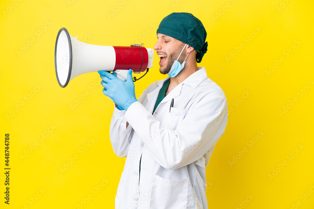 Surgeon blonde man in green uniform isolated on yellow background shouting through a megaphone Surgeon blonde man in green uniform isolated on yellow background shouting through a megaphone