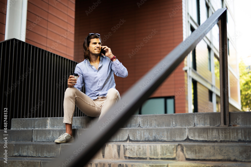 Fototapeta premium Young man sitting on the stairs drinking coffee. Handsome businessman using the phone while drinking coffee