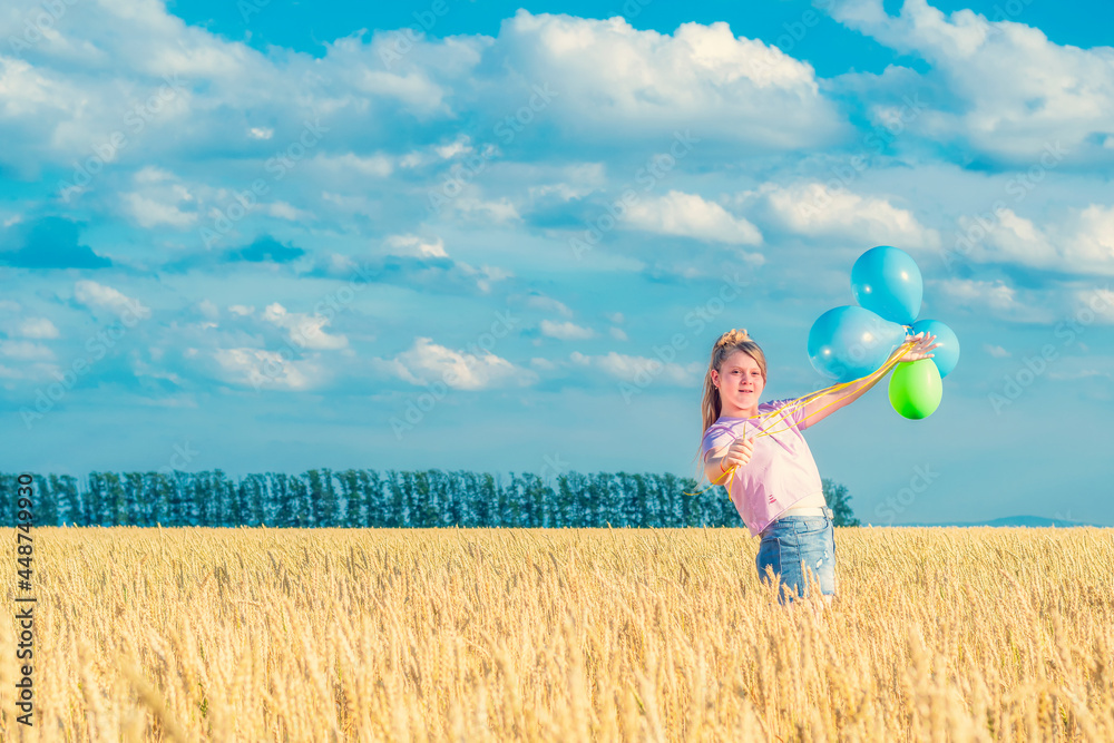 A young girl with colorful balloons in her hands is standing among a yellow wheat field against a blue cloudy sky. Looks into the camera. A young girl with colorful balloons in her hands is standing among a yellow wheat field against a blue cloudy sky. Looks into the camera.