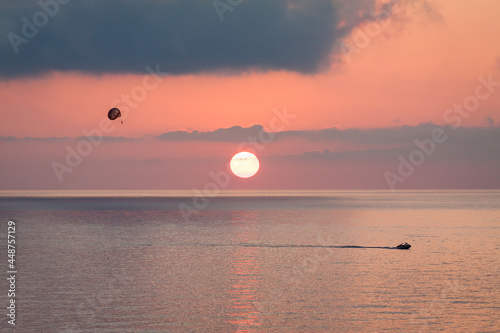 Parasailing behind a boat at sunset over the sea with the sun goes down. Silhouette of a person with parachute against evening sky with clouds. Beautiful seascape.