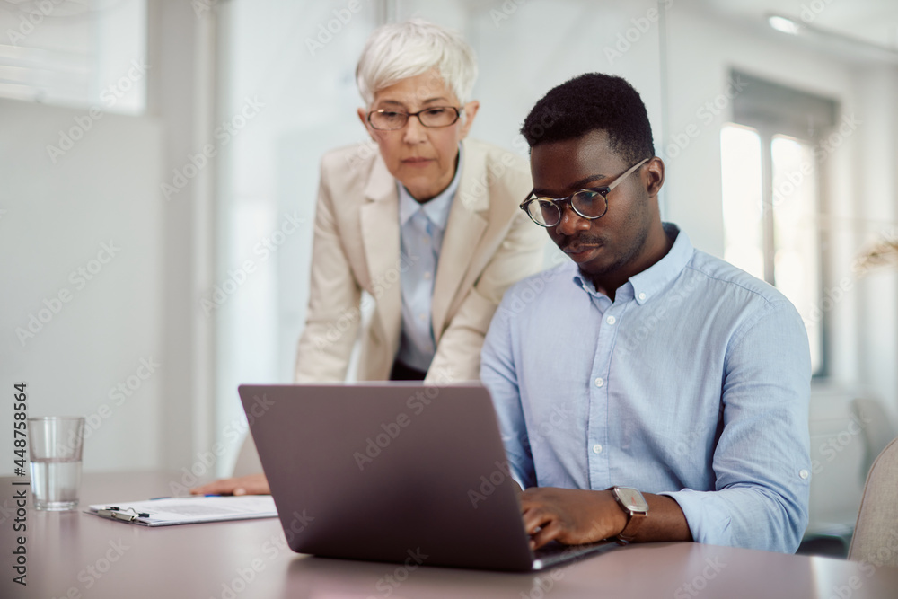 African American businessman works on laptop while mature mentor supervises him in office. African American businessman works on laptop while mature mentor supervises him in office.
