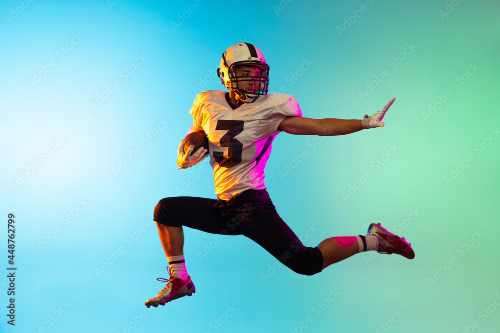 Portrait of American football player catching ball in jump isolated on blue studio background in neon light.