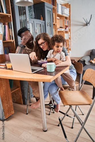 Woman feeding with breakfast her lovely multiracial child while sitting in front of laptop