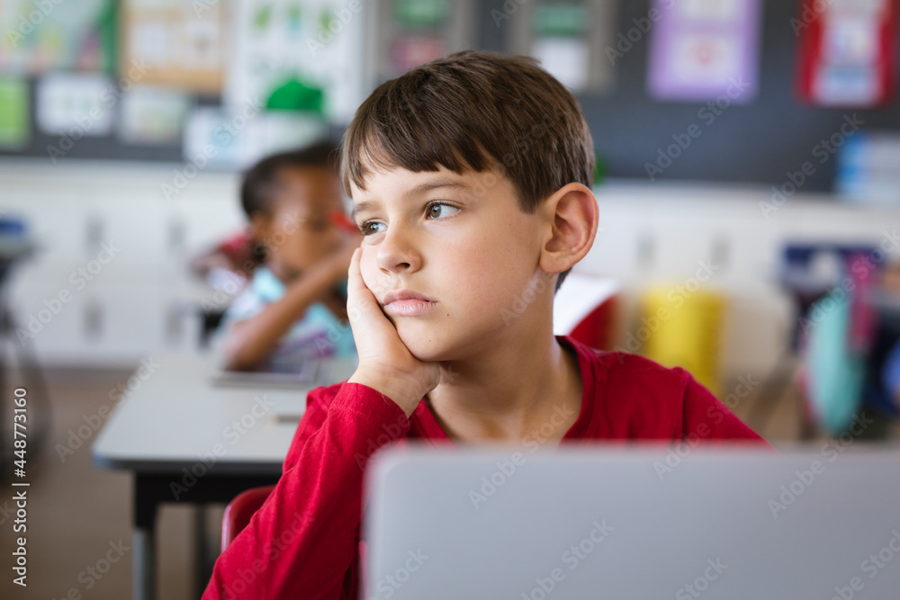 Sad caucasian boy with laptop sitting on his desk in the class at elementary school
