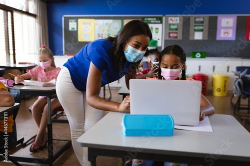 African american female teacher wearing face mask teaching a girl to use laptop at elementary school
