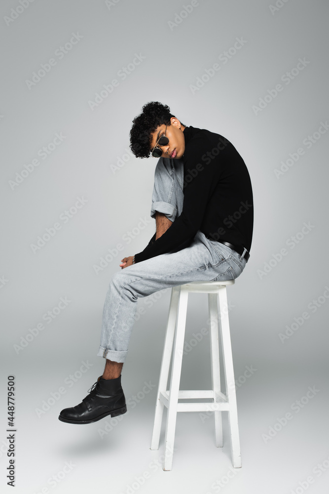 brunette african american man in jeans, turtleneck and sunglasses sitting on high stool on grey background brunette african american man in jeans, turtleneck and sunglasses sitting on high stool on grey background