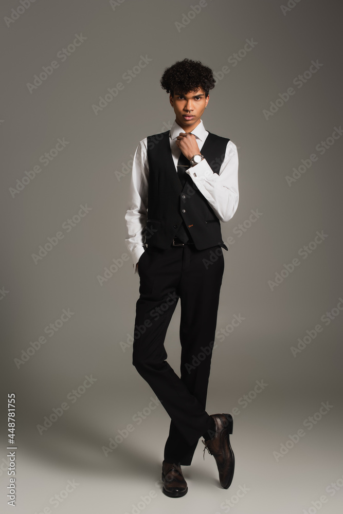 elegant african american man adjusting black tie while posing with hand in pocket on grey background elegant african american man adjusting black tie while posing with hand in pocket on grey background