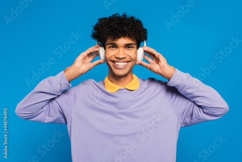happy african american man adjusting wireless headphones isolated on blue