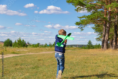 A six-year-old preschooler boy in a blue jacket launches a toy plane in a field against a blue sky with clouds on a summer day. The bright sun is shining. Scenery