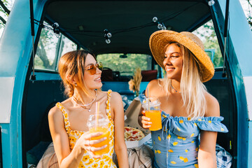 Two attractive cheerful women drinking lemonade near van, enjoying summer vibes in road trip