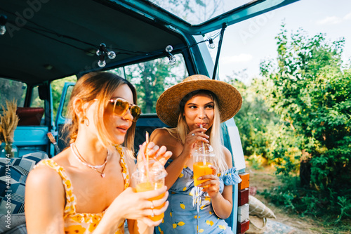 Two attractive cheerful women drinking lemonade near van, enjoying summer vibes in road trip