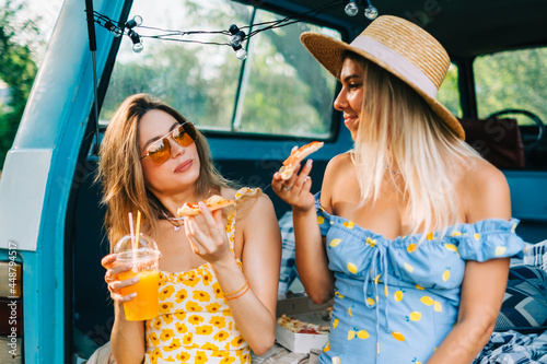Two attractive cheerful women drinking lemonade near van and eating pizza, enjoying summer vibes in road trip