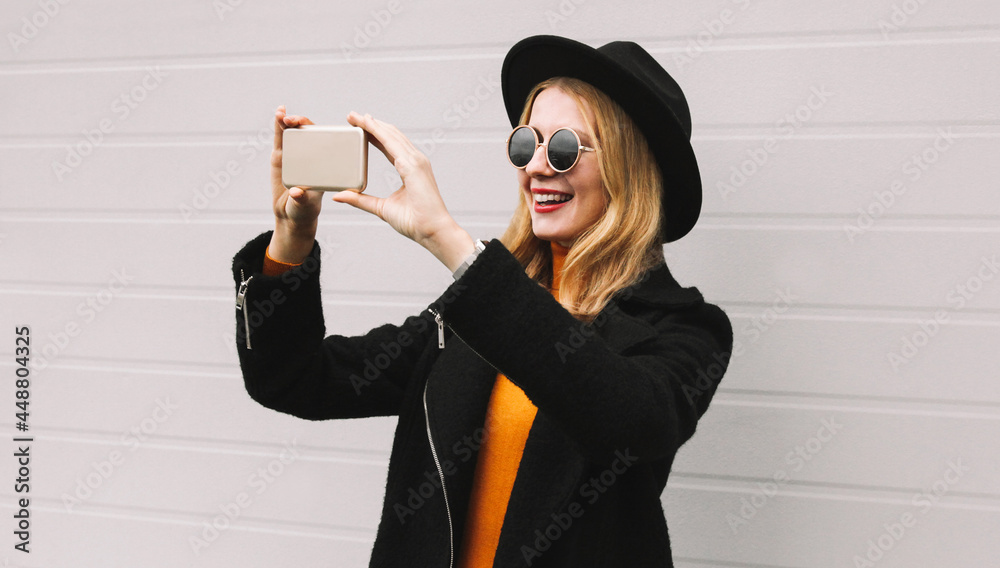 Portrait of stylish cheerful laughing young woman taking selfie by smartphone wearing a black coat, round hat on gray background Portrait of stylish cheerful laughing young woman taking selfie by smartphone wearing a black coat, round hat on gray background