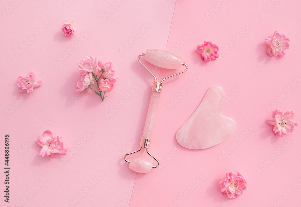 Facial massage kit for home spa. Face roller  and gua sha massager made from rose quartz  on pink pastel background with rose buds. Natural treatment concept.