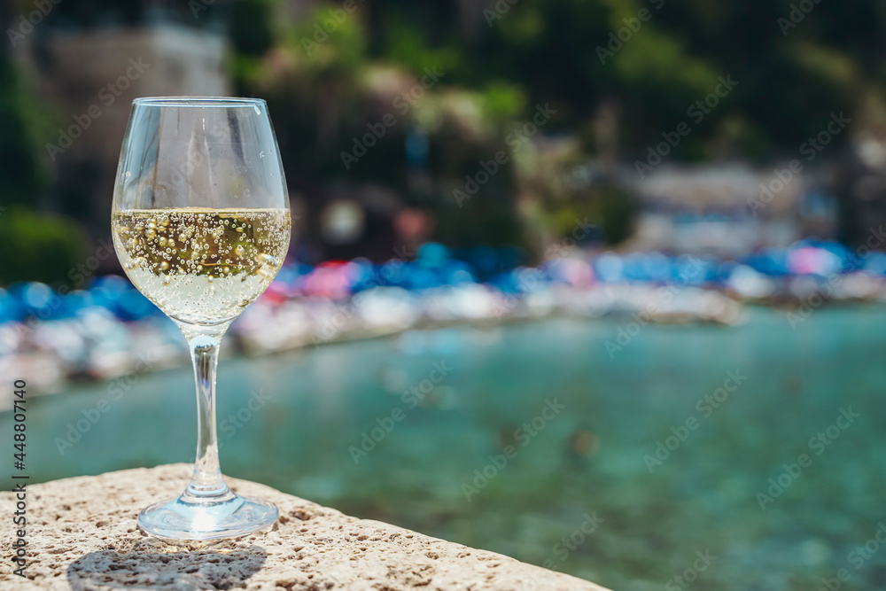 Glass of white wine against the background of the Mediterranean beach and the sea in a tourist town in the summer under sunlight. Summer, travel, lifestyle, relaxation, and enjoyment concept. Glass of white wine against the background of the Mediterranean beach and the sea in a tourist town in the summer under sunlight. Summer, travel, lifestyle, relaxation, and enjoyment concept.