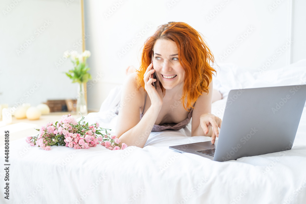 attractive ginger woman middle age in pajamas lying in bed using phone and laptop attractive ginger woman middle age in pajamas lying in bed using phone and laptop