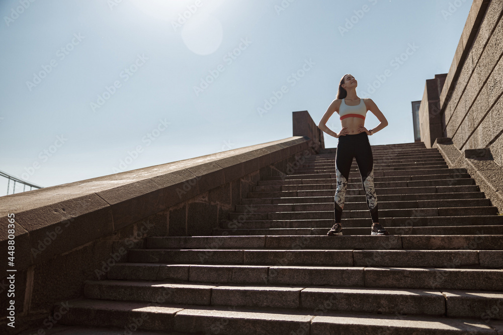 Woman in tracksuit with hands on waist stands on stone stairs on sunny summer day