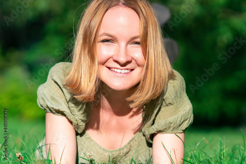 Slika na platnu Portrait of happy beautiful pretty cheerful girl, young positive red head ginger woman lying on grass in summer sunny park in dress, looking at camera, smiling