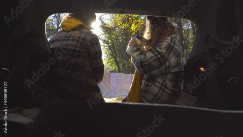 Long haired man and curly woman in checkered coat watching map and choosing direction in open trunk of hatchback car on road in autumn forest, backside view from salon