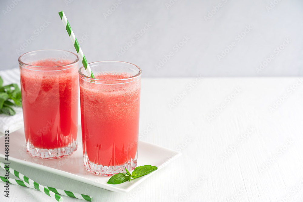 Two glasses of refreshing cold juice or smoothie made of ripe red watermelon served with paper straw and basil leaf on white wooden table in hot summer season. Image with copy space, horizontal Two glasses of refreshing cold juice or smoothie made of ripe red watermelon served with paper straw and basil leaf on white wooden table in hot summer season. Image with copy space, horizontal