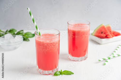 Watermelon refreshing cold red juice served in two drinking glasses with paper straw and green basil leaves on white wooden background with fruit slices as dessert and ice cubes summer season.