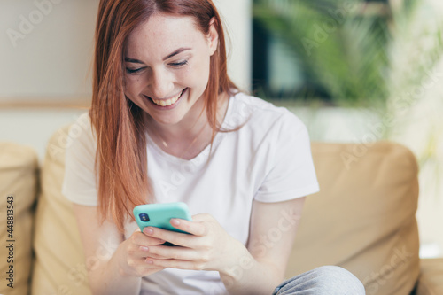 Young woman at home receiving positive emotions winning and good news while reading phone while sitting on sofa
