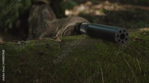 Close-up view of military marksman in camouflage putting out sniper rifle muzzle with suppressor on wooden log, looking through scope of optical sight, taking aim in dense forest at daybreak photo