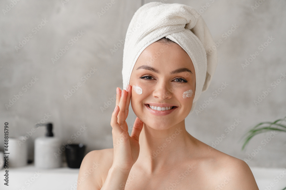 Young beautiful woman in bath towel smiling applying face body cream on her face for rejuvenation soft moisturizing effect. Spa beauty treatment concept. Body and skin care Young beautiful woman in bath towel smiling applying face body cream on her face for rejuvenation soft moisturizing effect. Spa beauty treatment concept. Body and skin care