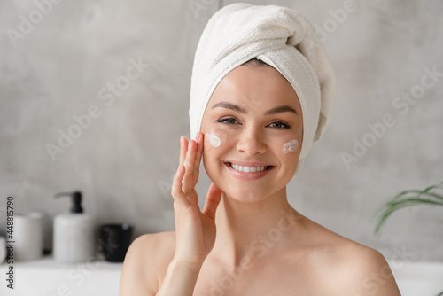 Young beautiful woman in bath towel smiling applying face body cream on her face for rejuvenation soft moisturizing effect. Spa beauty treatment concept. Body and skin care