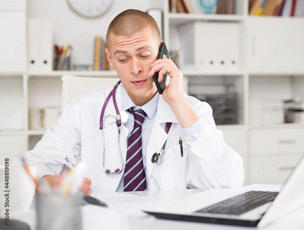 Young european doctor talking on phone and making notes in modern clinic office
