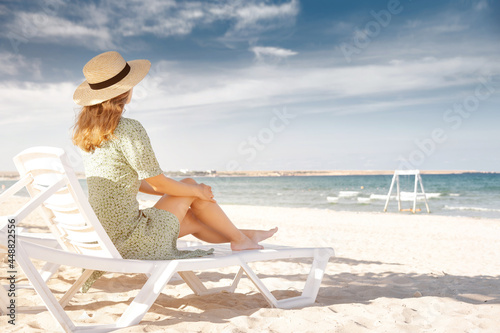 Woman on a beautiful beach in a green dress and a straw hat under wooden palm trees, relaxing on a sun lounger. Tropical island paradise, summer vacation or vacation.