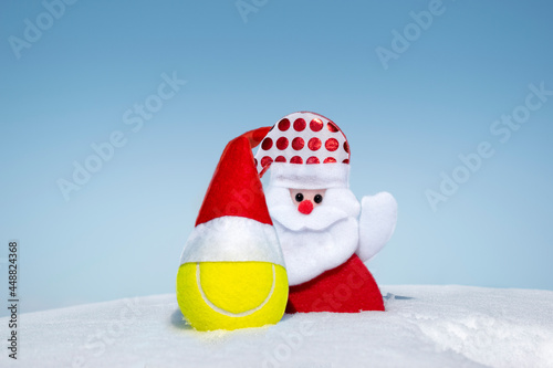 Santa Claus and tennis ball in red santa hat on white snow on winter day. Merry christmas and happy New Year greeting card, creative christmas composition, background, copy space